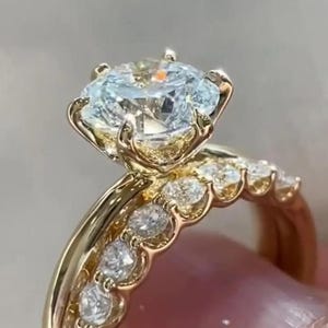 Può includere: Un set di anelli in oro con una grande pietra centrale a taglio rotondo sostenuta da sei griffe, accompagnata da una fede abbinata decorata con una fila di pietre più piccole a taglio rotondo. Il metallo ha una finitura gialla lucida.