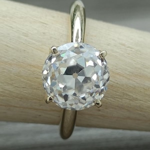 May include: A solitaire ring featuring a round, faceted clear gemstone set in a polished gold band. The stone is secured by six prongs, showcasing a brilliant cut with a distinct culet at the center, mounted on a smooth, rounded metal band.