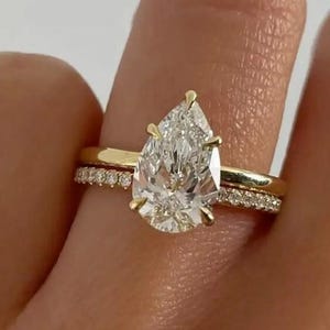 May include: A gold ring set featuring a pear-shaped diamond center stone. The ring is set on a gold band, with a smaller band of pave diamonds. The ring is shown on a finger.