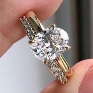 May include: A gold engagement ring featuring a large oval-cut diamond, paired with a band set with smaller diamonds. The rings are held up, highlighting the brilliance of the gemstones. The design is classic and elegant.