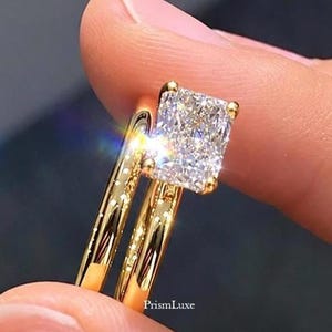 May include: A gold ring set featuring a radiant-cut clear gemstone with a high-shine finish. The band is polished yellow gold, and the setting includes the text PrismLuxe. The stone reflects light with a prismatic effect, showcasing clear facets.