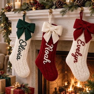 Op de afbeelding: Drie hangende kerstsokken van kabelgebreid materiaal. De linker is crème met een donkergroene strik en de naam Olivia. De middelste is rood met een crèmekleurige strik en de naam Noah. De rechter is crème met een donkerrode strik en de naam Evelyn.