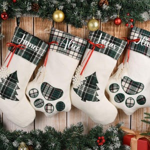 May include: Four personalized Christmas stockings with names James, Alice, Lucas, and Olivia. Each stocking is cream-colored with a green and black plaid cuff and accents. The stockings are decorated with a Christmas tree and paw print designs.