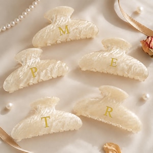 May include: Five off-white hair claw clips with a marbled texture, each featuring a single gold letter printed in the centre. The letters displayed on the clips are M, P, E, T, and R. These hair accessories have a curved, scalloped edge design.