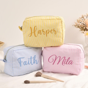 Può includere: Tre trousse per cosmetici personalizzate in giallo, azzurro e rosa, ciascuna con un nome ricamato in un colore complementare. Le borse sono realizzate in un tessuto simile al seersucker e sono dotate di cerniera.