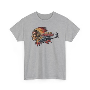 Op de afbeelding: Grijs t-shirt met een afbeelding van een Indiaanse chef met een hoofdtooi, een helikopter en de tekst "AH-64 APACHE". Het ontwerp is in rood, geel, zwart en wit.