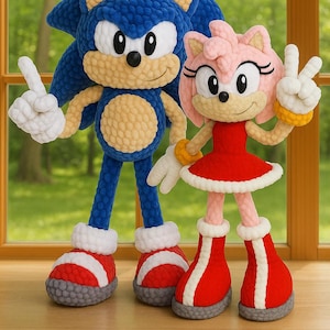 Könnte beinhalten: Gehäkelte Sonic the Hedgehog und Amy Rose Puppen. Sonic ist blau mit weißen Handschuhen und roten Schuhen. Amy Rose ist rosa mit einem roten Kleid und Schuhen. Beide Puppen stehen vor einem Fenster mit grünem Hintergrund.
