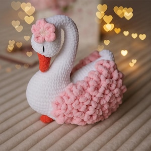 Puede incluir: Un cisne de ganchillo blanco y rosa hecho a mano. El cisne tiene un cuerpo blanco, un cuello curvado y una flor rosa en la cabeza. Las alas del cisne son de color rosa esponjoso, y tiene un pico y patas de color naranja. El cisne es un artículo decorativo.