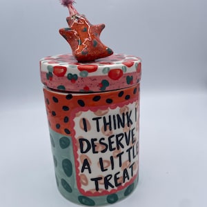 May include: A cylindrical ceramic jar with a lid and a star-shaped topper. The jar is decorated with red, green, and black polka dots and the text "I THINK I DESERVE A LITTLE TREAT". The topper has a pink flower.