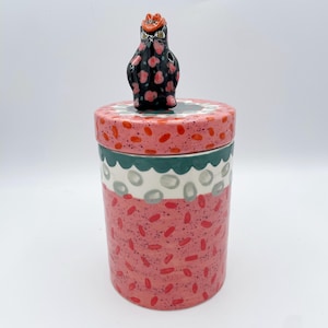 May include: A ceramic jar with a lid, featuring a whimsical design. The jar is primarily pink with red speckles, and the lid is adorned with a black owl figurine with pink floral accents. The jar's lid has a green and white scalloped border.