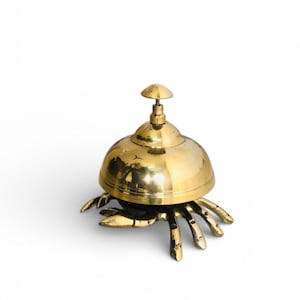 Può includere: Una campana a forma di granchio in ottone con una parte superiore a forma di cupola e una piccola maniglia. La campana ha sei zampe di granchio ed è posta su uno sfondo bianco. La campana è un oggetto decorativo.