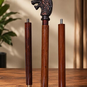 Peut inclure: Une canne de marche en trois parties avec un fût en bois brun foncé et une poignée métallique en forme de tête de loup. La poignée présente une finition bronze sombre et patinée. Chaque segment en bois possède un raccord fileté.