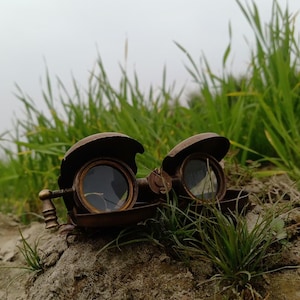 May include: Antique brass-colored steampunk goggles with round lenses and protective covers. The goggles are resting on a sandy surface with green grass in the background, evoking a vintage aesthetic.