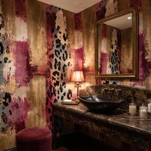 May include: A bathroom interior featuring wallpaper with a bold abstract pattern of gold, deep magenta, and black leopard spots. A dark marble vanity holds a black vessel sink, a gold-framed mirror, a table lamp, and glass containers.