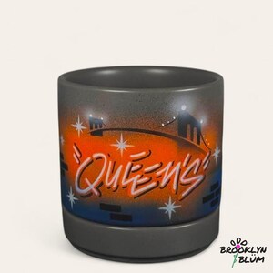 Pode incluir: Caneca cilíndrica de cerâmica com base e borda cinzentas. A caneca apresenta um design em estilo grafite com a palavra "QUEENS" em branco e laranja, contra um fundo de ponte e luzes da cidade. A caneca tem um logotipo Brooklyn Blum.