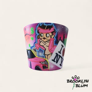 Puede incluir: Una maceta de cerámica pintada a mano con una mujer de dibujos animados con atuendo rosa, un coche rosa y el texto "I NY". La maceta tiene un paisaje urbano y símbolos de dólar. El logotipo de Brooklyn Blum está en la parte inferior.