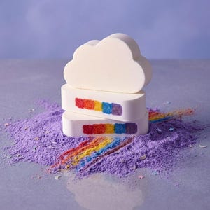 Puede incluir: Tres bombas de baño blancas, una en forma de nube y dos rectangulares, con detalles de arcoíris. Descansan sobre un lecho de polvo morado con un rastro de arcoíris. El fondo es de color púrpura suave.