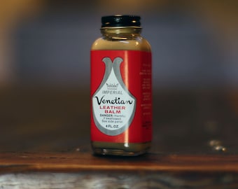 May include: A small, clear glass bottle with a red label that says "Imperial Venetian Leather Balm". The bottle is sitting on a wooden surface.