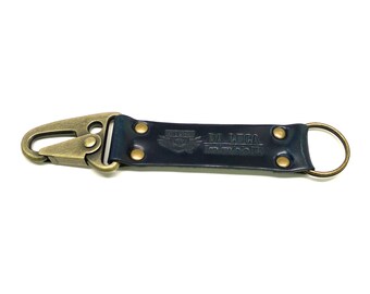 May include: A black leather keyring with a brass hook and a brass ring. The keyring has the text "DA LUCA" and a logo embossed on it.