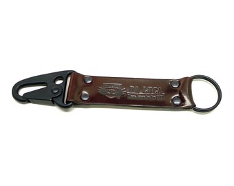 May include: A brown leather key chain with a black metal clip and a black metal ring. The keychain has the text "DA LUCA" and a logo with a bird with wings spread.