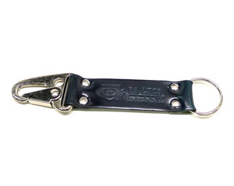 May include: A black leather keyring with a silver metal clasp and key ring. The keyring features the text "DA LUCA Handmade in USA" and a logo. The keyring is designed to attach to a belt loop or bag.