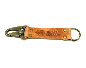 May include: A brown leather key chain with a brass hook and a brass ring. The key chain has the text "DA LUCA Handmade in USA" and a logo with a bird and the text "DA LUCA" embossed on it.