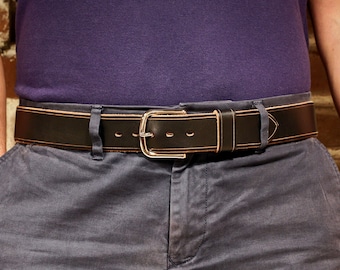 May include: A brown leather belt with a silver buckle. The belt is worn around the waist of a person wearing a purple shirt and grey trousers.