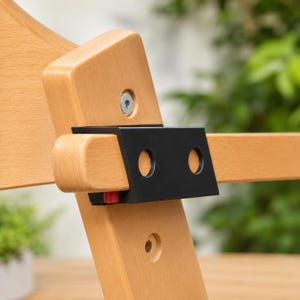 Könnte beinhalten: Ein helles Holzmöbelteil mit einer schwarzen Metallhalterung, die mit einer silbernen Schraube befestigt ist. Die Halterung hat zwei kreisförmige Aussparungen und einen kleinen roten Kunststoff-Entriegelungsmechanismus an der Unterseite.