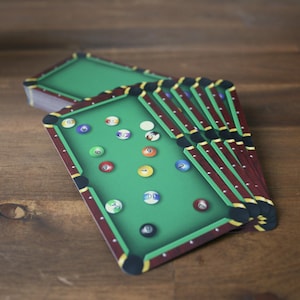 Könnte beinhalten: Billiard-Themen-Untersetzer mit grünem Tischdesign und nummerierten Kugeln. Die Untersetzer haben einen bordeauxfarbenen Rand mit gelben Akzenten und liegen auf einer Holzoberfläche.