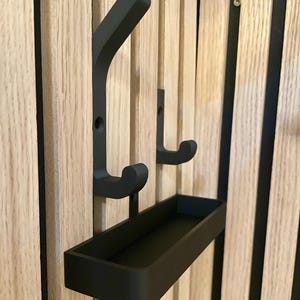 Könnte beinhalten: Ein schwarzer Wandorganizer mit zwei Haken und einer kleinen Ablage. Der Organizer ist auf einer hellen Holzplatte mit vertikalen Rillen montiert. Die Haken sind gebogen und die Ablage ist rechteckig.