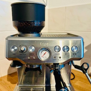 Könnte beinhalten: Eine Sage Espressomaschine aus Edelstahl mit integriertem Bohnenmahlwerk. Die Maschine hat mehrere Knöpfe und ein Manometer. Das schwarze Bohnenmahlwerk hat einen transparenten Behälter mit Kaffeebohnen. Auf einer Holzoberfläche.