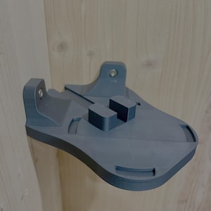 Könnte beinhalten: Eine graue, 3D-gedruckte Wandhalterung mit geschwungenem Design. Sie verfügt über zwei rechteckige Blöcke und eine Halterung mit einer Schraube. Die Halterung ist für die Befestigung an einer Ecke der Wand konzipiert.
