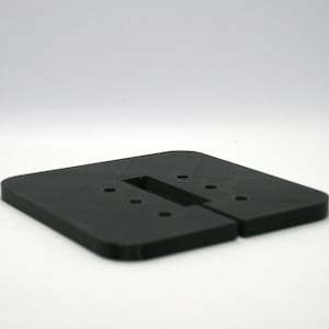 Könnte beinhalten: Eine flache, quadratische schwarze Grundplatte mit abgerundeten Ecken, einem rechteckigen Ausschnitt in der Mitte und einer Reihe kleiner kreisförmiger Löcher, die symmetrisch auf der Oberfläche angeordnet sind. Das Objekt besteht aus festem, mattem schwarzem Material.