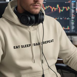 Puede incluir: Una sudadera con capucha beige con el texto "EAT SLEEP TRADE REPEAT" en negro. La persona lleva auriculares negros y está sentada frente a múltiples pantallas de ordenador que muestran gráficos bursátiles. Se ven un teclado y un ratón.