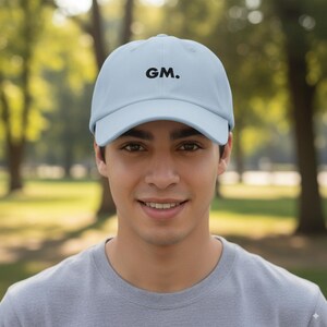 Op de afbeelding: Lichtblauwe baseballcap met de geborduurde tekst "GM." in zwart. De cap heeft een gebogen klep en een klassiek zes-panelen ontwerp. De hoed is gemaakt van een zachte, ademende stof.