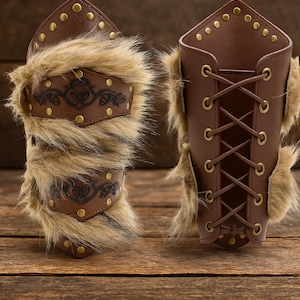 May include: Two brown leather arm guards with fur trim and decorative metal studs. One arm guard has a decorative leather panel with an embossed design. The other arm guard has a lace-up closure.