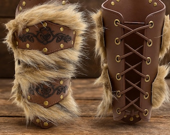 May include: Two brown leather arm guards with fur trim and decorative metal studs. One arm guard has a decorative leather panel with an embossed design. The other arm guard has a lace-up closure.