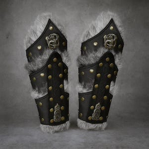 Könnte beinhalten: Paar schwarze Leder-Armschützer mit Messingnieten und dekorativen Metall-Emblemen. Die Armschützer haben einen grauen Pelzbesatz und sind im mittelalterlichen oder Fantasy-Stil gestaltet. Das Leder ist mit Nähten und Nieten versehen.