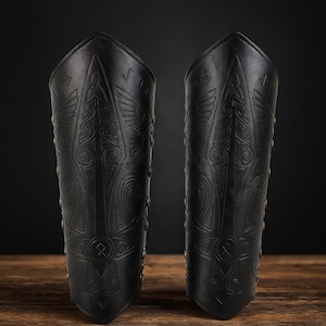 May include: Pair of black leather arm guards with intricate embossed designs. The arm guards feature detailed patterns, including stylized wings and geometric shapes. Rivets line the edges, adding to the rugged aesthetic. The background is dark, highlighting the texture and craftsmanship.