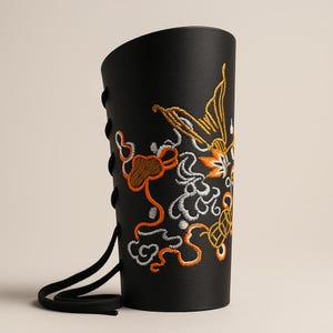 Puede incluir: Brazalete de cuero negro con intrincados bordados en naranja, blanco y dorado. El brazalete presenta un cierre con cordones y un borde superior curvado. El diseño bordado incluye patrones florales y abstractos.