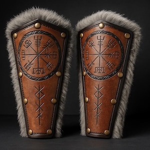 Puede incluir: Par de protectores de brazo de cuero marrón con un diseño circular en relieve y tachuelas de latón. Los protectores de brazo presentan un ribete de piel y símbolos grabados. El diseño recuerda a la armadura vikinga o medieval.