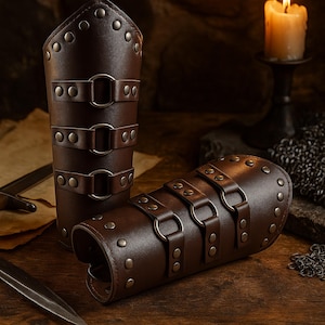 Peut inclure: Deux protège-bras en cuir marron avec rivets et boucles en métal. Les protège-bras sont cylindriques, avec trois sangles et boucles. L'un est droit, l'autre est couché. Une bougie et une épée sont en arrière-plan.