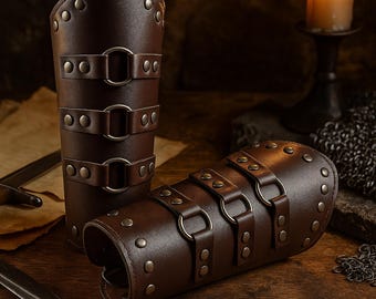 May include: Two brown leather arm guards with metal rivets and buckles. The arm guards are cylindrical, with three straps and buckles. One is upright, and the other is lying down. A candle and a sword are in the background.