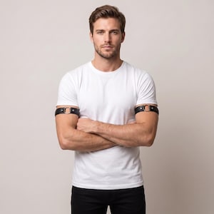 Può includere: Una coppia di bracciali in pelle nera con un anello metallico circolare centrale e finiture color argento. Le fasce sono fissate con piccoli rivetti e si indossano intorno alla parte superiore delle braccia, creando un accessorio dal design netto.