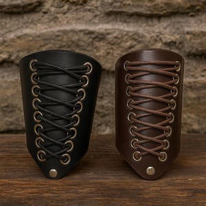 Puede incluir: Se muestran dos brazaletes de cuero, uno negro y otro marrón. Cada brazalete presenta un diseño con cordones con ojales metálicos y un cierre de botón. Los brazaletes están colocados sobre una superficie de madera y un fondo de piedra texturizada.