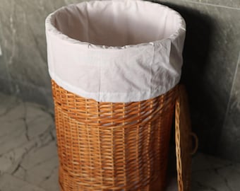 May include: A round, woven wicker laundry hamper with a light-colored fabric liner. The hamper is a natural brown color and has a matching lid. The hamper is open, revealing the white fabric liner. The hamper is next to a gray wall.