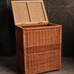 May include: A rectangular, open-top wicker laundry hamper with a hinged lid. The hamper is made of woven light brown wicker and has a light-colored wooden lid. The hamper is on a gray tiled floor.