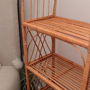May include: A light brown wicker shelf with three levels and an arched top. The shelves are made of horizontal slats, and the sides feature decorative woven patterns. The shelf is designed for storage and display.