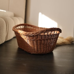 May include: A brown wicker basket with an oval shape. The basket has a woven design and a braided rim. It is sitting on a dark wooden floor.