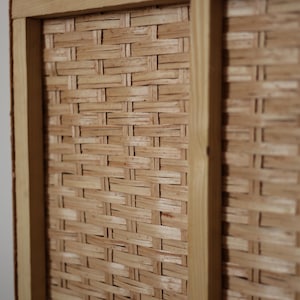 Pode incluir: Close-up de uma cesta de vime tecida em tons castanhos claros. A cesta tem formato retangular, com uma moldura de madeira clara. A trama intrincada do vime &eacute; claramente vis&iacute;vel, criando uma superf&iacute;cie texturizada.
